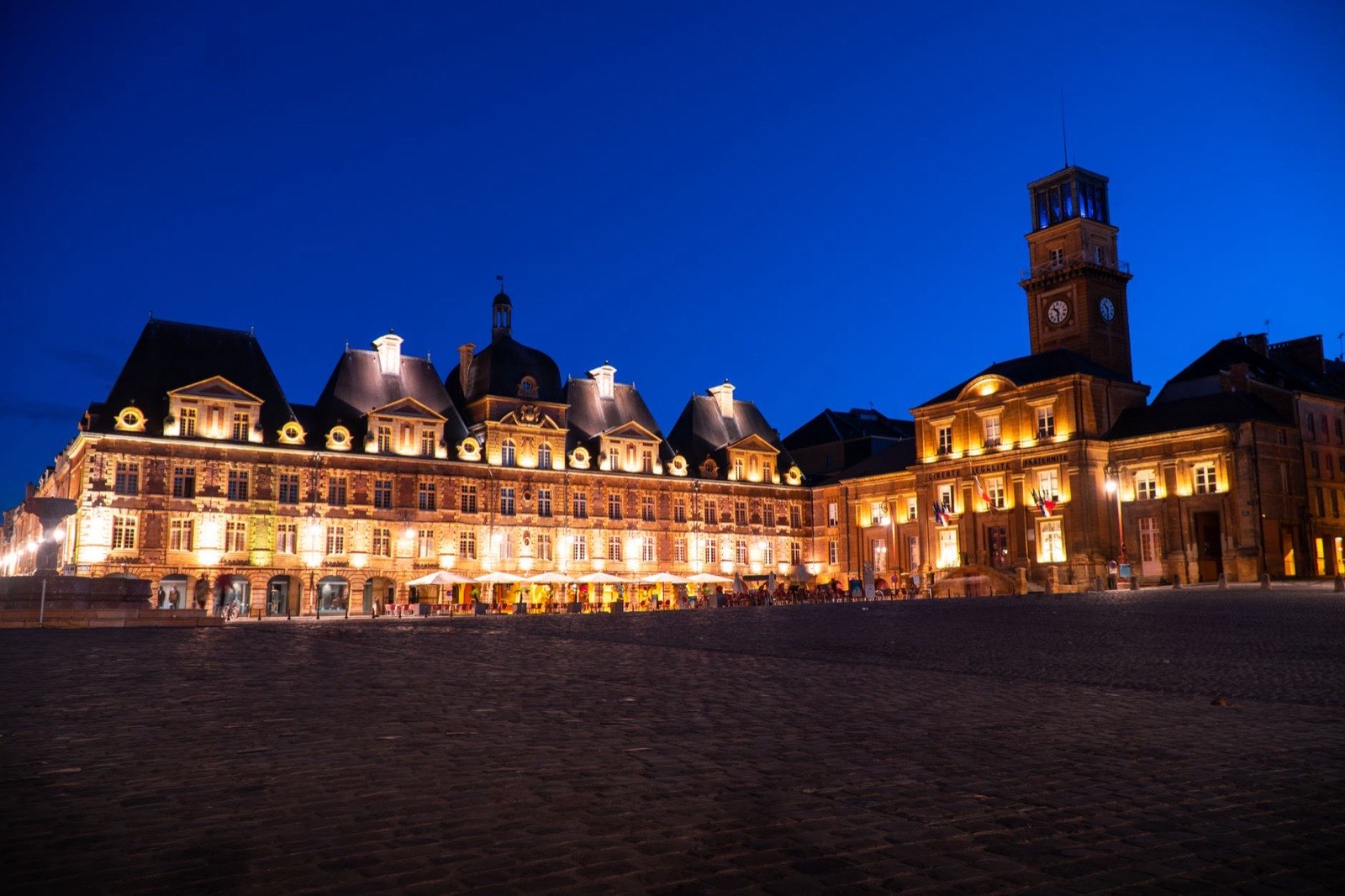 Place d’une ville française à l’heure bleue, hôtel de ville éclairé portant la devise républicaine et pavoisé aux trois couleurs.